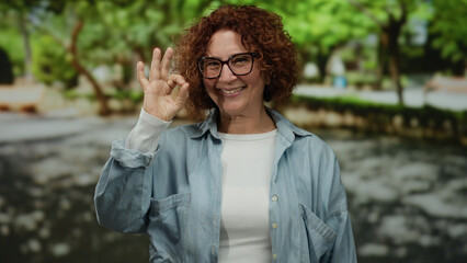 Woman smiling in a park, wearing glasses and casual clothes, gesturing thumbs up with a bright...