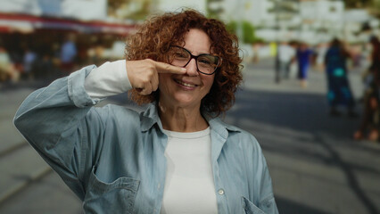 Fototapeta premium Woman smiling on a sunny city street pointing to her nose wearing glasses and a casual shirt with a blurred urban backdrop bustling with people.