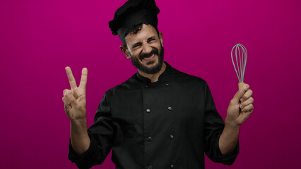 Hispanic bearded man chef smiling in black uniform holding whisk on a vibrant pink background,...