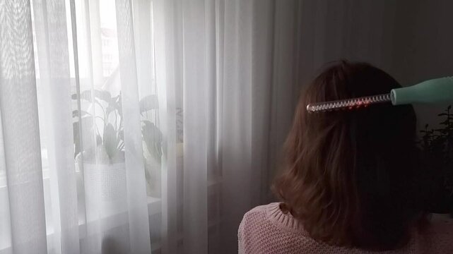 A woman runs a pulse massager through her hair, view from the back . Hair care at home with the Darsonval pulse massager.