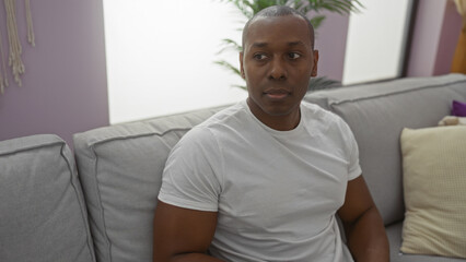 Young african american man sits in living room with modern decor, relaxed on a grey sofa in a cozy...