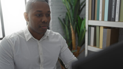 Young man working in modern office setting, focused on computer with bookshelf and plants in background, conveying a professional and serene environment.