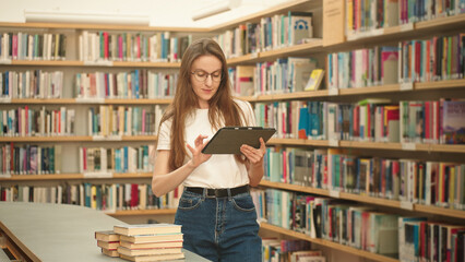 University student using a tablet to search for books in the library. Modern academic research, project research, digital tools, and exam preparation. Woman reading or studying academic knowledge goal