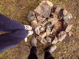 Fire pit made of rocks on a grass and people feet next to it. The fire place is small and full or dirt and ash and the rocks are scattered around it. Camping and cooking food in wild concept.