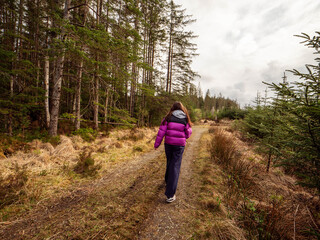 A girl in a purple jacket and dark pants walks on a dirt narrow road in a forest park. The road is...