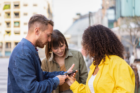 Three young friends interacting and laughing while looking at a smartphone together on a city street. They enjoy a digital moment in an urban environment. Modern lifestyle and technology concept.