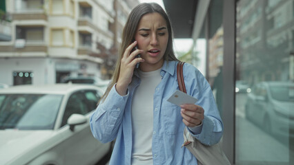 Obraz premium Woman speaking on phone holding card on busy street wearing blue shirt near cars buildings outdoor.