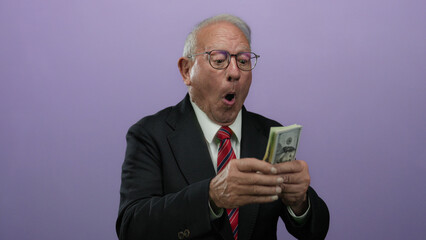 Senior man excitedly counting us dollar banknotes against a purple background, showcasing business success and financial abundance.