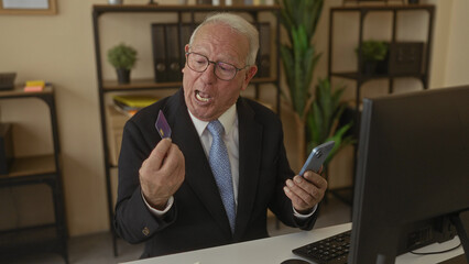 Senior man in suit sits in office holding credit card and smartphone smiling at computer in a modern workplace setting.