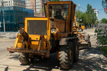 Grader operates on city street, smoothing and leveling asphalt for new pavement. Construction equipment, urban surroundings, and ongoing roadwork highlight infrastructure development and maintenance