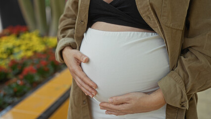 Pregnant woman gently holding baby bump outdoors with colorful flowers in background, capturing...