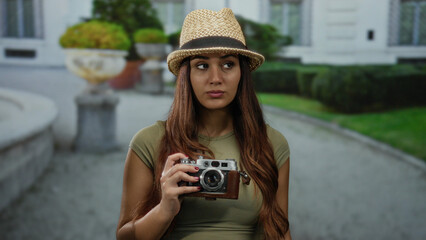 Woman tourist holding camera outdoors on a street wearing hat and looking around near a building...