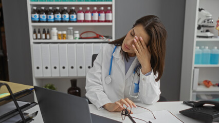 Woman doctor sitting stressed in hospital clinic workplace with medical equipment in background appears exhausted with head resting on hand during workday.