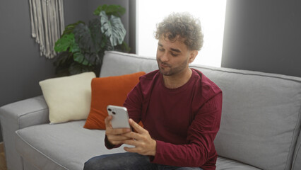Young hispanic man in casual attire using smartphone on cozy sofa in stylish living room interior