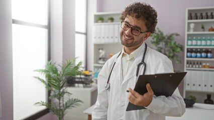 Handsome hispanic male doctor smiling in clinic room holding clipboard at workplace with bright windows and healthcare equipment in background