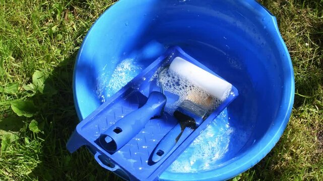 Painting tools soak in blue basin with soapy water on green grass after using water-based varnish, cleaning process in sunlight.