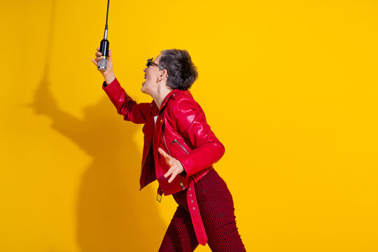 Energetic senior woman singing passionately while holding a microphone wearing stylish fashion against a vibrant yellow background