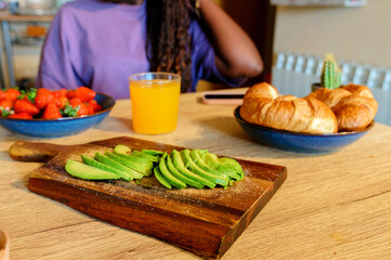 Fresh avocado slices seasoned with salt on a wooden cutting board, accompanied by croissants, strawberries, and orange juice, create a vibrant and nutritious breakfast spread