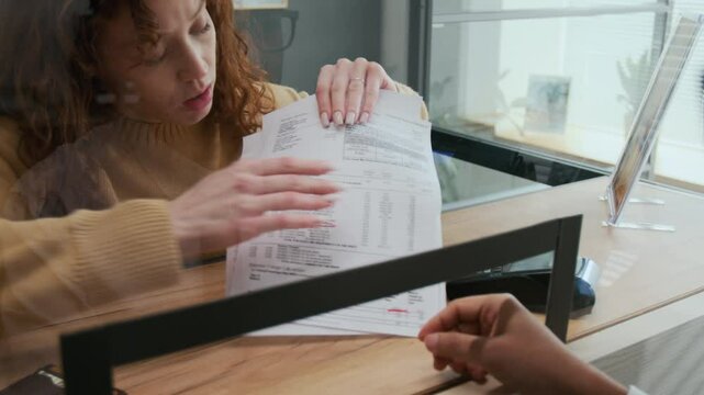 Confused red-haired woman asking bank teller to help her with document issue at counter in modern office