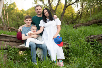 Fototapeta premium Family posing for photo in park. Two children with parents. Teenage boy, baby, mom and dad sitting on log outdoors in summer. 