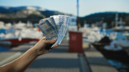 Man holds hungarian forint banknotes by a seaside port with boats in the background, emphasizing outdoor, water setting and financial travel concept.