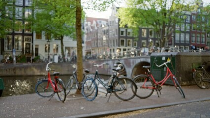 Fototapeta premium Blurred bicycles line the canal in amsterdam under green trees with historic buildings softly defocused in the bustling netherlands urban scene.