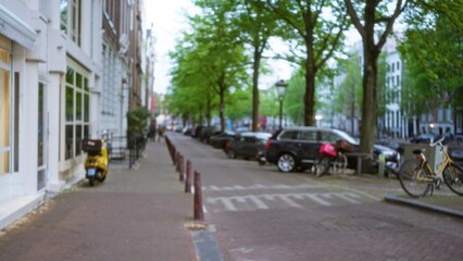 Obraz premium Blurry city street in amsterdam with bicycles and parked cars lined along the sidewalk under green trees, capturing the essence of urban life in the netherlands with a defocused view.