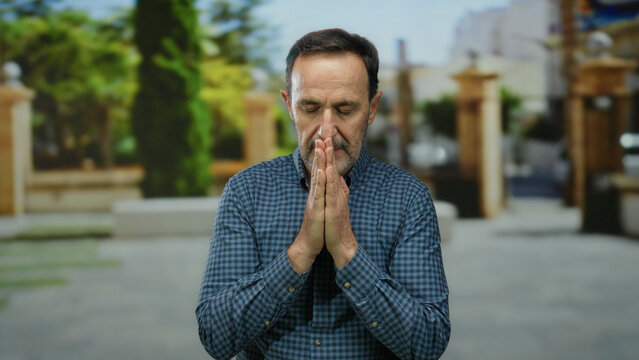 Elderly man meditating outdoors in an urban park setting with eyes closed and hands in prayer pose wearing a checkered shirt under sunny weather surrounded by greenery.