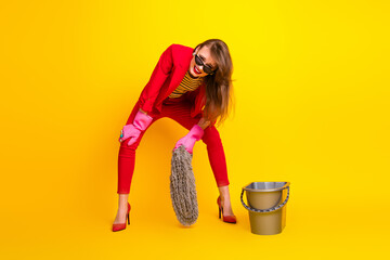 Stylish young woman in red suit and pink gloves having fun cleaning with mop on bright yellow background