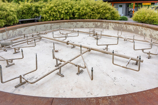 An empty fountain with a complex pipe system can be seen in the center of a well-kept park. The rusted pipes and chipped plaster at the edge of the pool indicate disuse or the need for renovation.