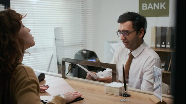 Over the shoulder shot of unrecognizable woman with red curly hair interacting with bank assistant through glass teller window in office