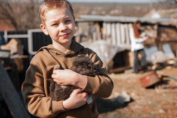 A young boy holds a black puppy in his arms while standing in a rustic backyard. The moment captures the connection between a child and his pet, with a countryside setting in the background.