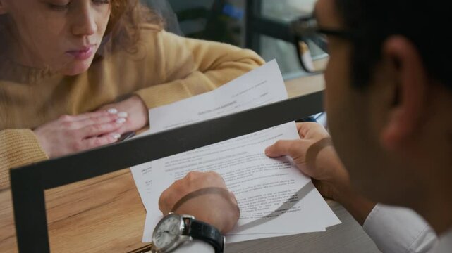 Close-up over the shoulder shot of unrecognizable male bank assistant giving instructions to red-haired woman across counter in office
