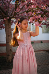 Young girl in a pink dress standing under blooming cherry blossom trees, placing a flower in her...