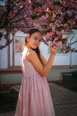 A young girl in a pink dress gently touches cherry blossoms while smiling under a blooming tree. A...