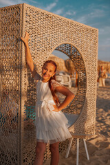 Smiling girl in a white summer dress posing by a stylish wicker screen on a sunny sandy beach with...