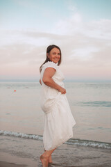 Happy woman in a white flowing dress enjoying a barefoot walk along the beach at sunset. Soft waves and pastel sky add a peaceful, carefree vacation vibe.