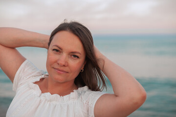 Portrait of a smiling woman in a white dress posing by the sea with her hands behind her head. Calm...