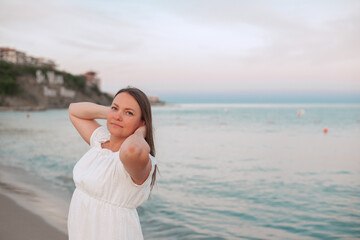 Relaxed woman in a white dress enjoying a calm evening on the beach. She holds her hands behind her...