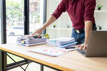 A person is sitting at a desk with a laptop and a stack of papers. They are working on a project and have a potted plant nearby
