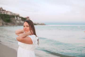 A woman in a white dress stands on a sandy beach near the sea, looking back with a calm smile....