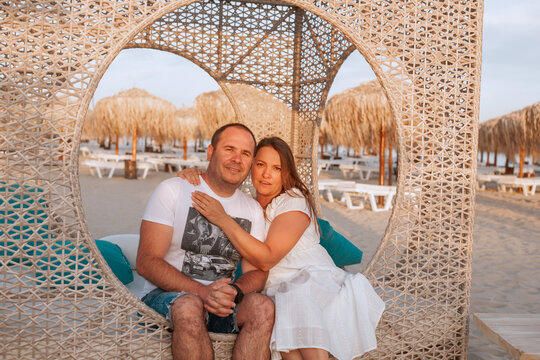 Couple sitting together in a cozy beach lounge at sunset. They are embracing each other warmly, surrounded by straw umbrellas and sandy beach. Romantic summer vacation mood.