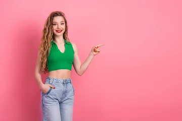 Gordijnen Smiling young woman pointing with hand on pink background, wearing green top and jeans, showcasing trendy fashion style © deagreez