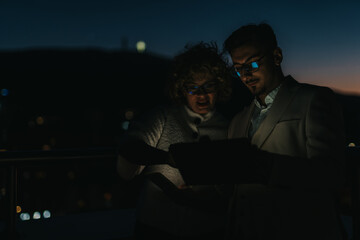 Two people collaborate over a project at night, utilizing technology under a dimly lit sky. The...