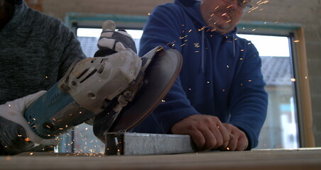 Worker cutting metal with angle grinder, sparks flying while another worker holds the metal steady,...