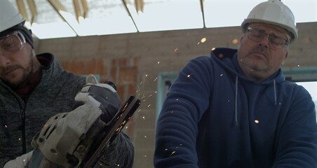 Obraz premium Two men in protective gear cutting metal with angle grinder, sparks illuminate construction site, teamwork and precision in super slow motion