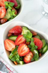 Fresh salad with strawberries and cucumbers in heart shaped bowl on white kitchen table. Top view.