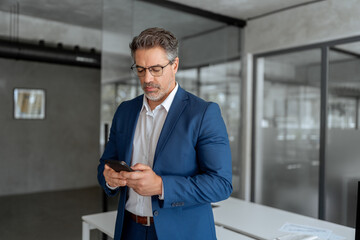 Middle eastern arab or latin senior business manager man ceo using cell phone mobile. Focused Indian mature businessman in suit and eyeglasses holding smartphone standing in office, working on device