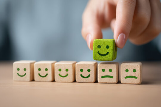 A hand places a green happy face block atop a row of wooden blocks with varying expressions, symbolizing the importance of customer satisfaction in achieving positive feedback