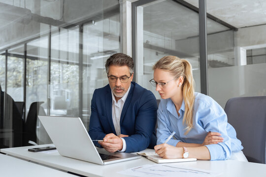 Two partners, team of professional business people working, discussing, writing notes at desk. Mature focused senior mentor leader man showing young woman manager project on laptop in modern office